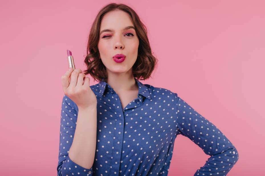 Mujer con blusa color azul y puntos blancos de frente sosteniendo un lipstick rosa, sobre las mejores marcas de lipstick para lucir labios perfectos
