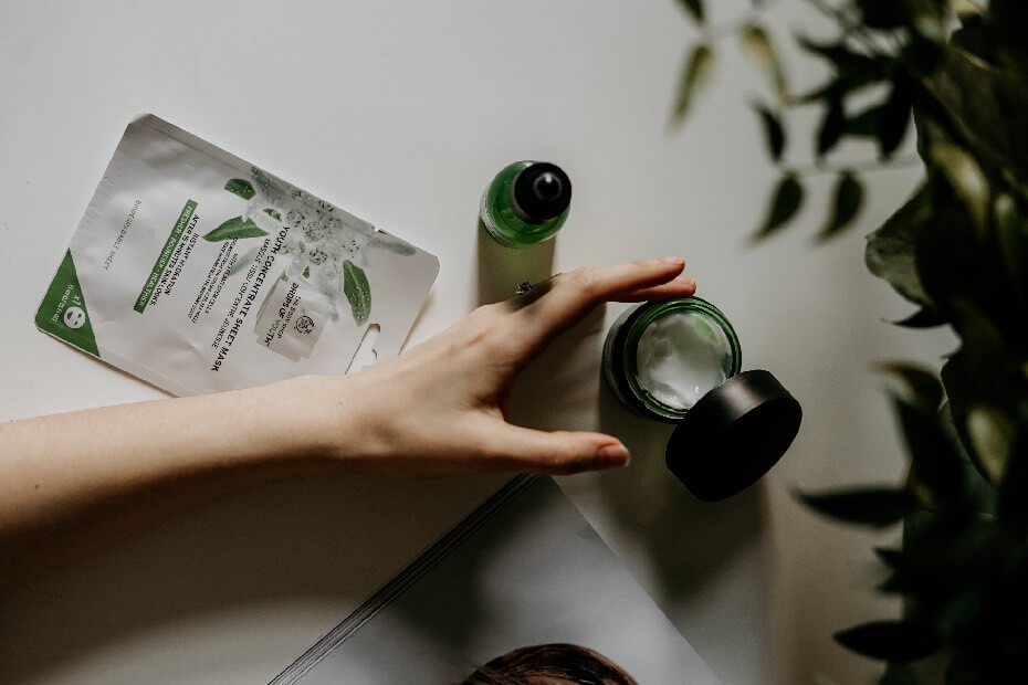 Una mano sujetando una crema y al lado una mascarilla, sobre productos de belleza que no pueden faltar en tu rutina diaria