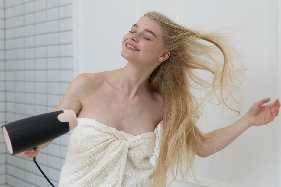 Mujer sonriendo y aplicándose aire de secadora en el cabello, sobre 5 tips para minimizar los daños causados por el uso excesivo de herramientas térmicas