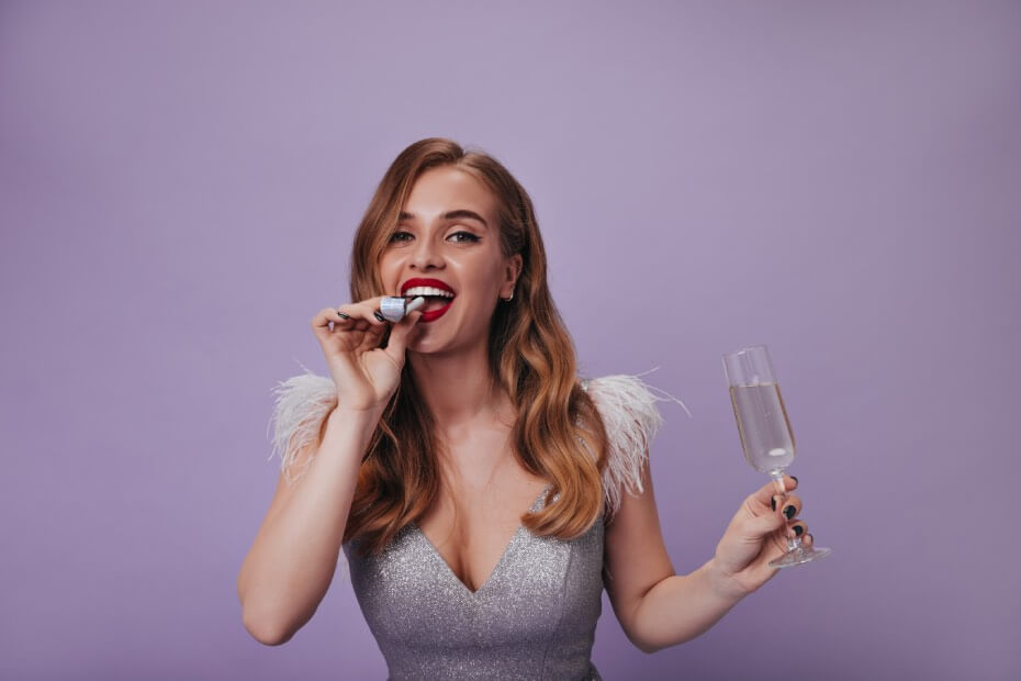 Mujer rubia atractiva con traje plateado sosteniendo su copa de champán sonriendo y disfrutando su fiesta de año nuevo 2024 sobre fondo púrpura